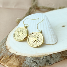 Load image into Gallery viewer, Gold compass-shaped earrings on a white shell with natural background