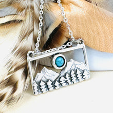 Load image into Gallery viewer, Necklace with a pendant featuring mountains and a blue gemstone, displayed on feathers.