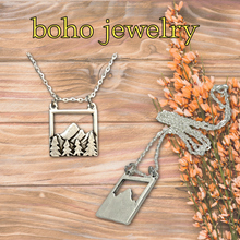 Load image into Gallery viewer, Boho jewelry necklace with mountain and tree design on a wooden surface with flowers.
